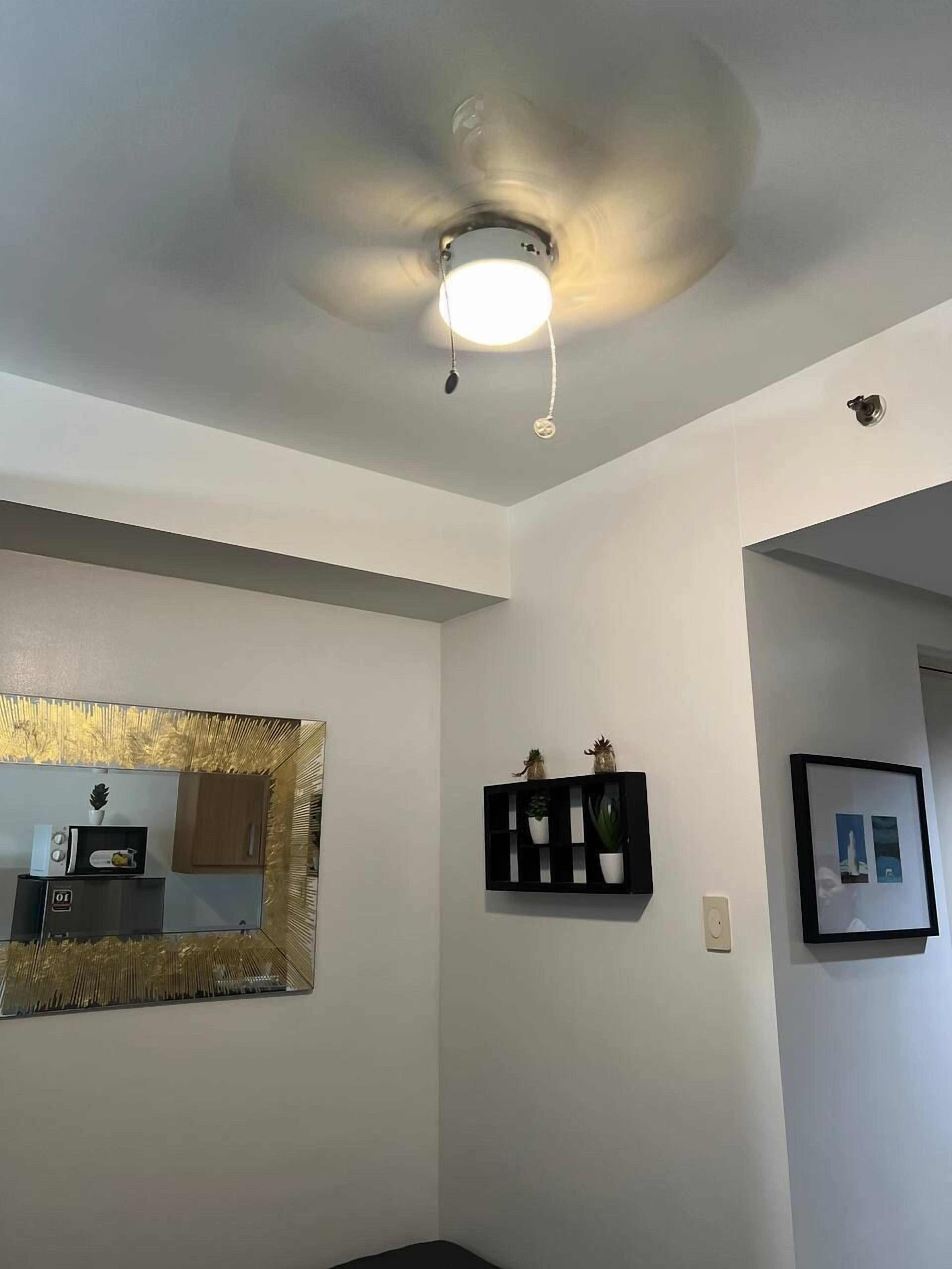Ein Zimmer mit Deckenventilator, spiegelt in der Wand, schwarzes Regal mit Dekoration und einem gerahmten Bild.