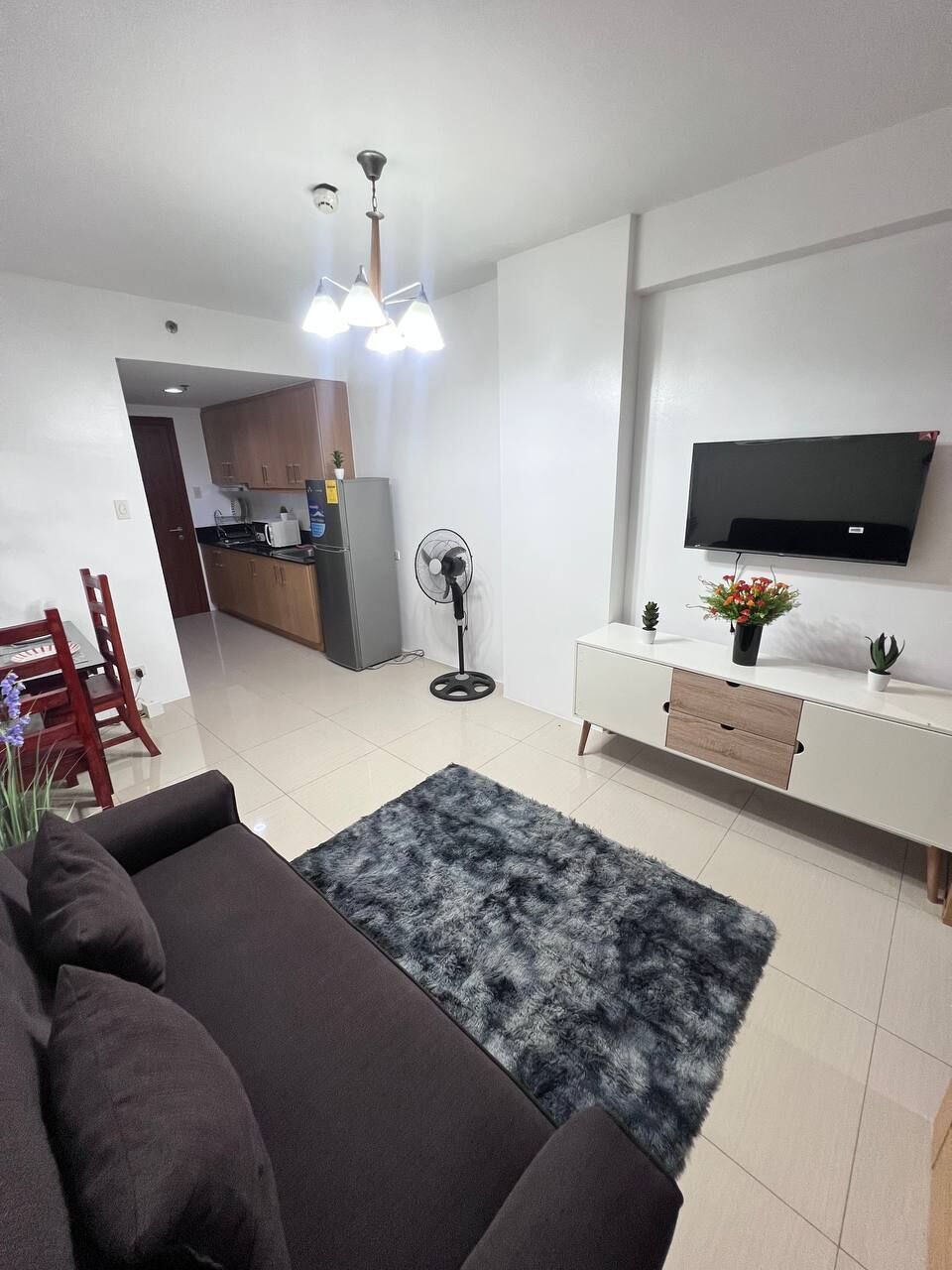 A modern living room with a sofa, a TV, a fan, and a kitchen in the background.