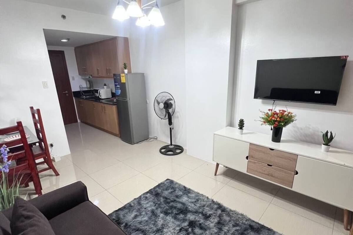 A modern living room with a sofa, a TV, a fan, and a kitchen in the background.