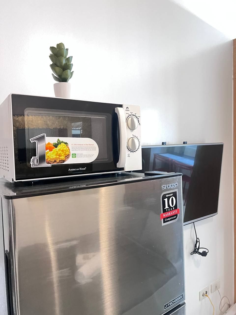 Una cocina compacta con un microondas sobre un refrigerador de acero inoxidable, una pequeña planta en maceta y un televisor de pantalla plana montado en la pared.