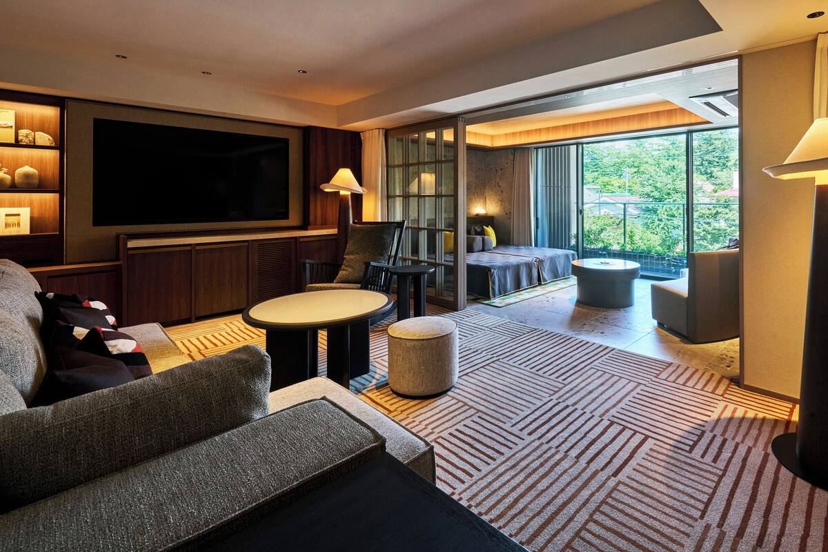 Ein modernes Hotelzimmer mit einem großen Flachbildfernseher, einem Sofa, einem Esstisch und einem Balkon mit Blick auf Grünflächen.