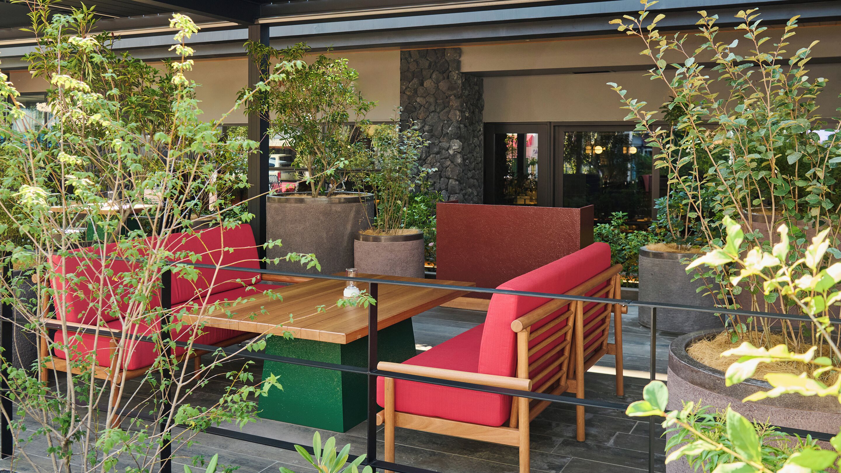 An outdoor seating area with red cushioned benches and a wooden table, surrounded by potted plants and a stone wall.