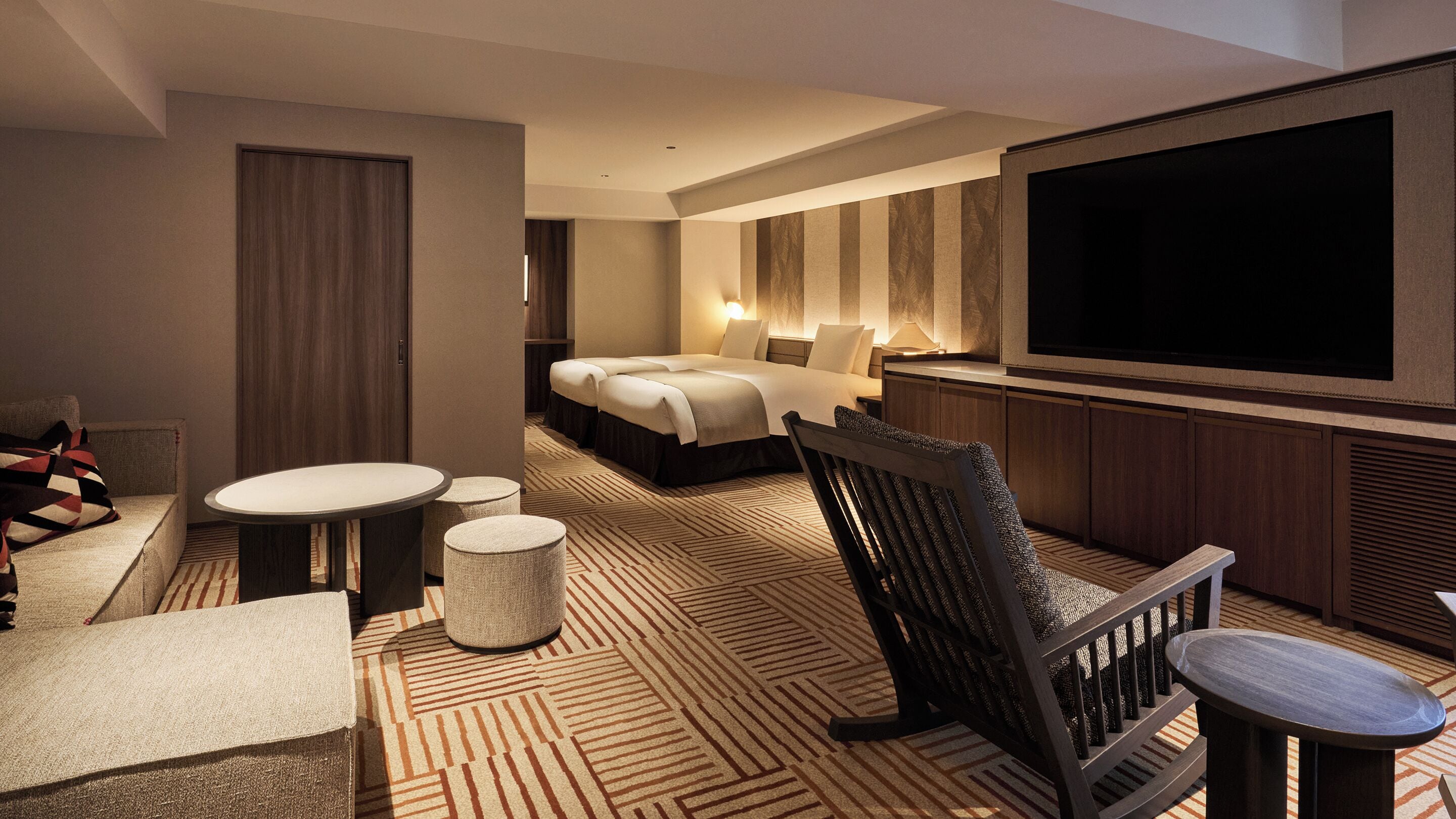A hotel room with a large bed, a TV, a seating area with a chair and ottoman, and a patterned carpet.