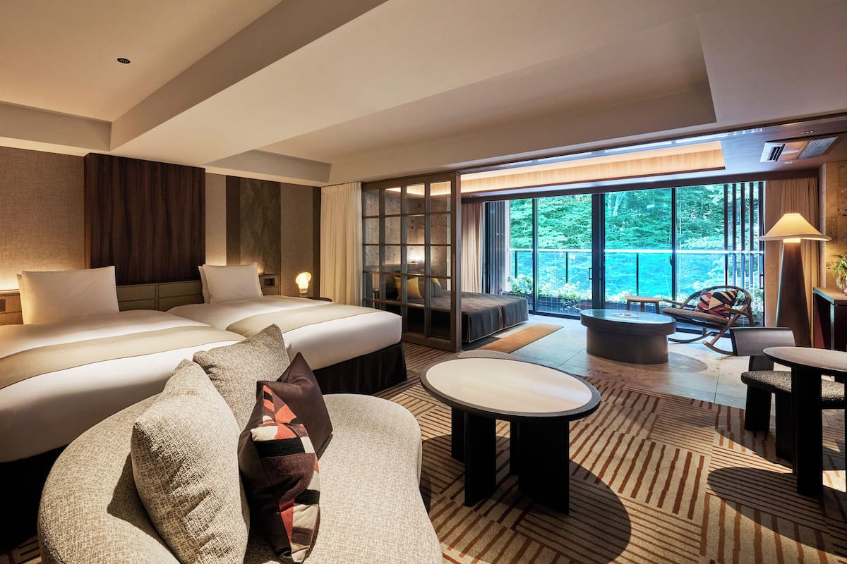 Ein Hotelzimmer mit einem großen Bett, zwei Nachttischen, einer Sitzecke mit Sofa und Sesseln, einem kleinen Tisch und Blick auf einen Balkon mit Pool.
