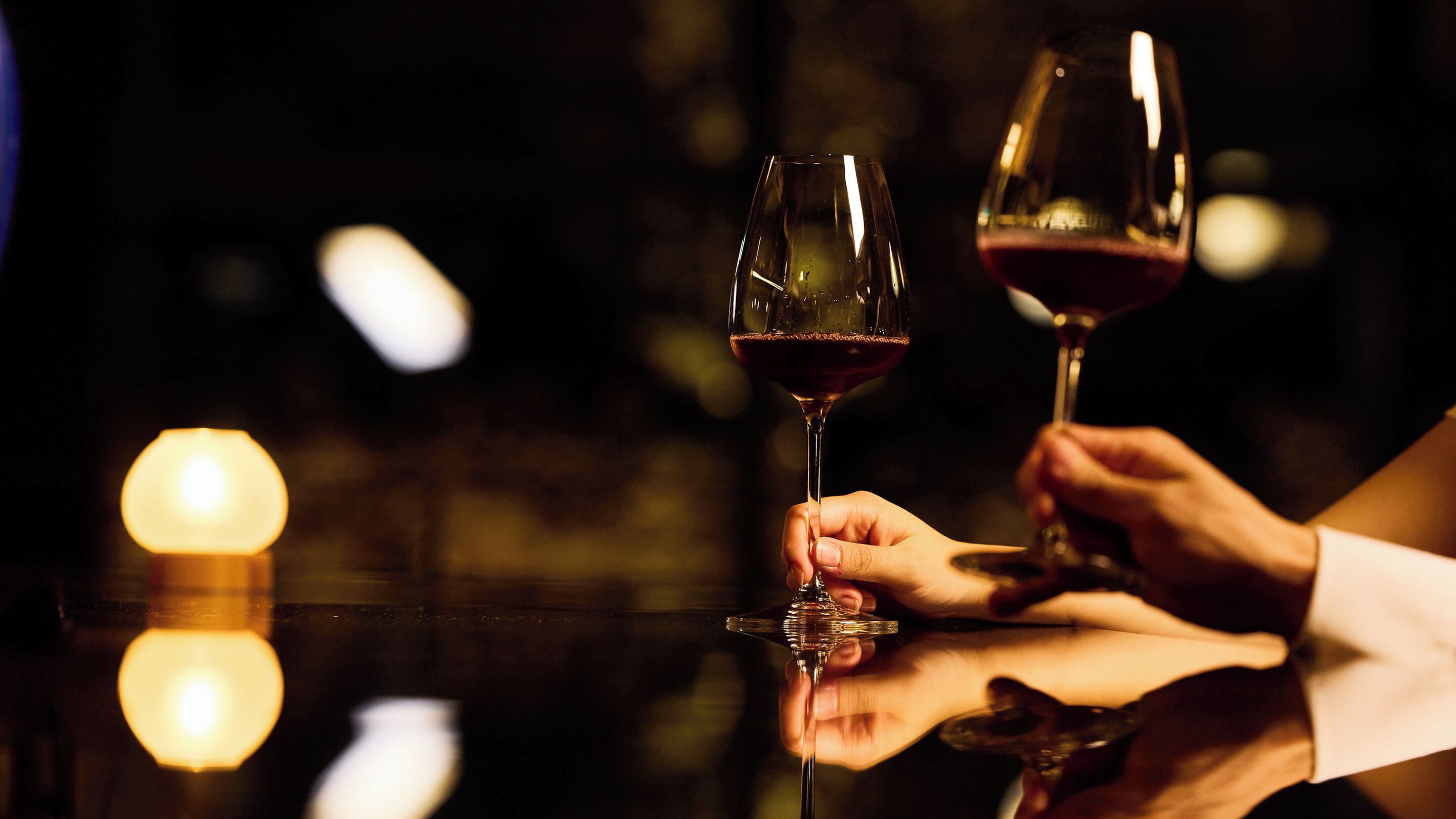 Zwei Hände halten Weingläser, eines mit Rotwein und eines mit Weißwein, vor dunklem Hintergrund mit unscharfen Lichtern.