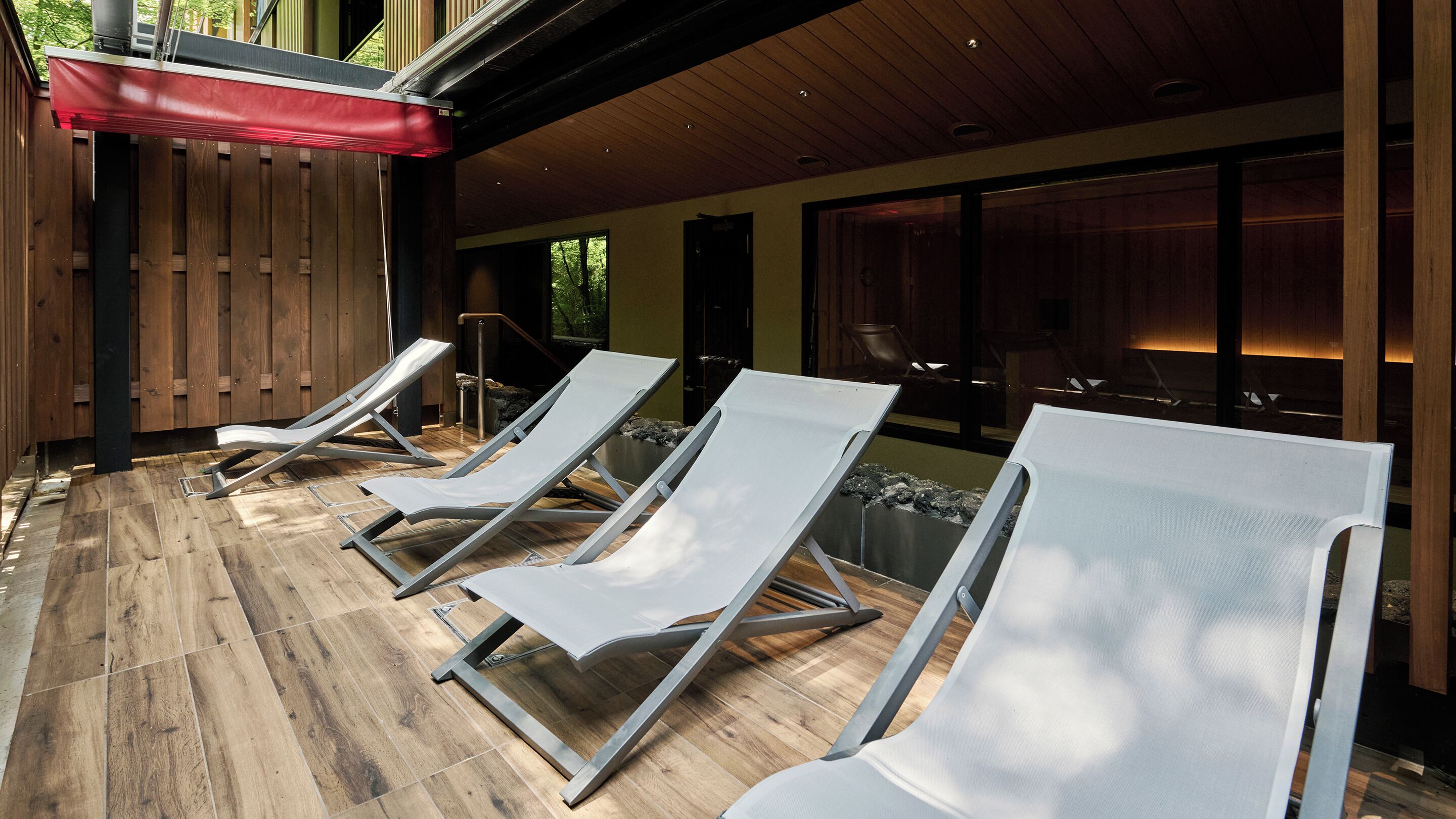 A covered outdoor area with four lounge chairs aligned in a row, wooden flooring, and a red awning.