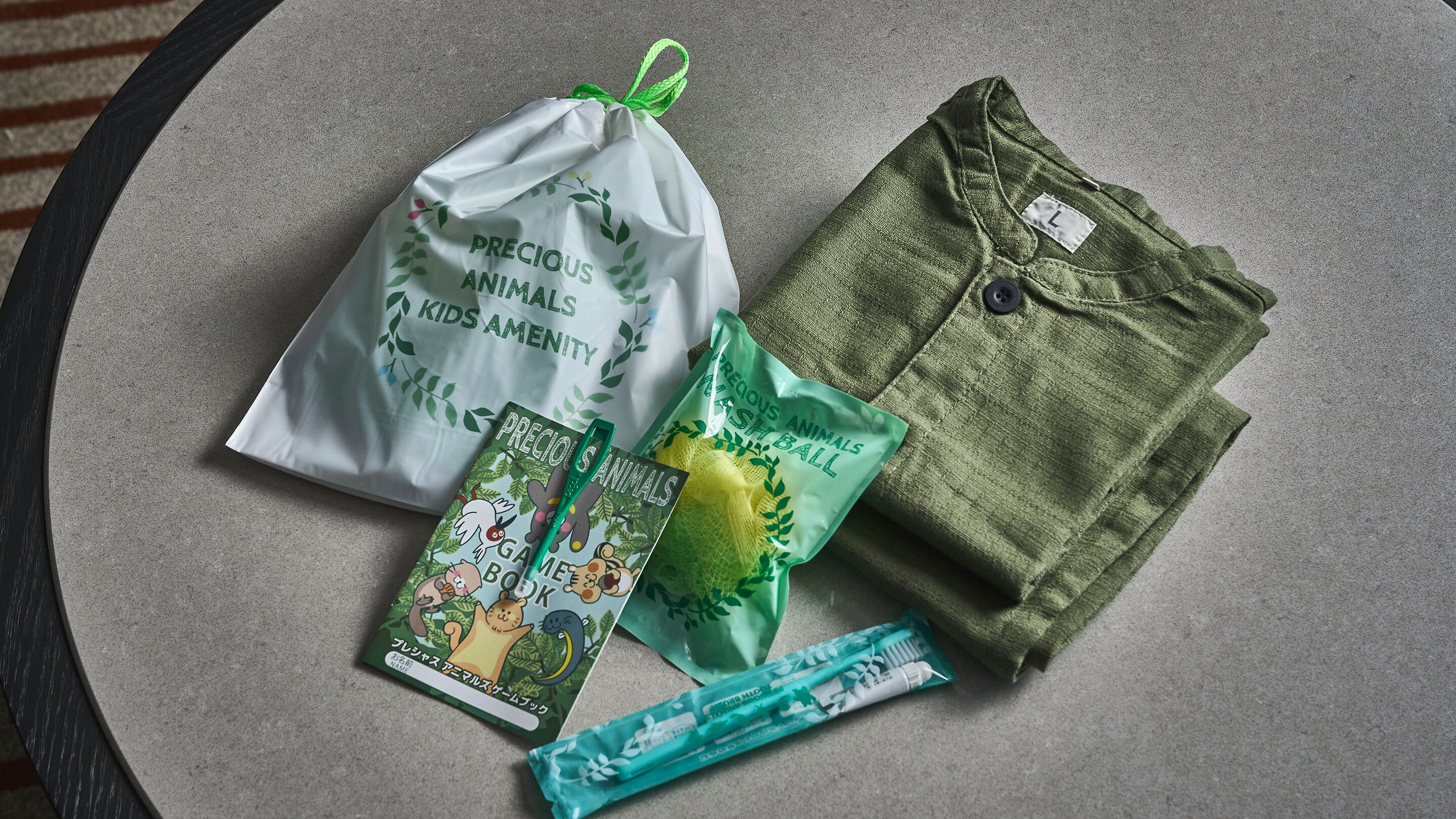 A white drawstring bag with 'PRECIOUS ANIMALS KIDS AMENITY' printed on it, a green mesh ball, a book titled 'GAME BOOK,' and a pair of green pants.