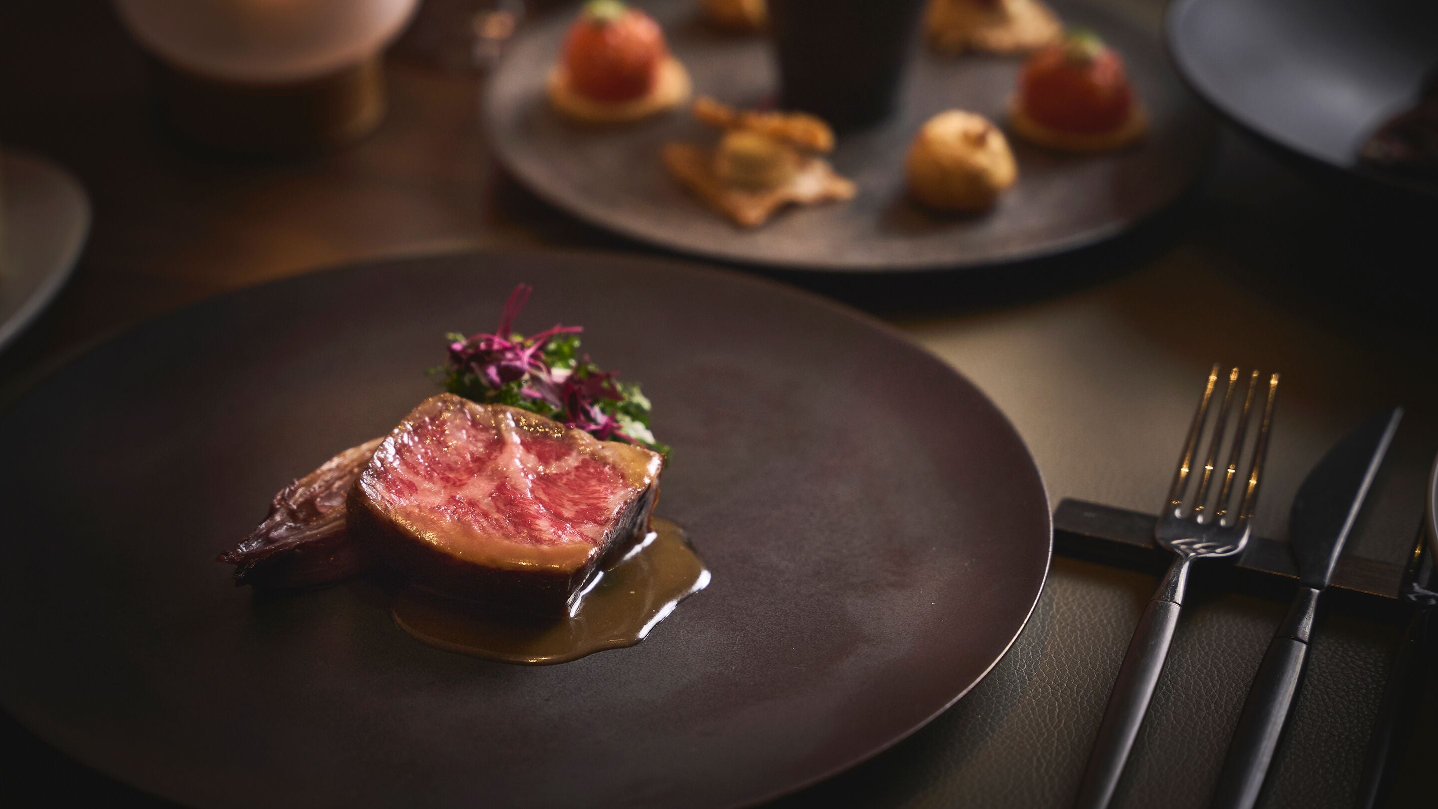 A plate with a steak and vegetables, a fork and knife on the right, and a blurred background with plates of food.