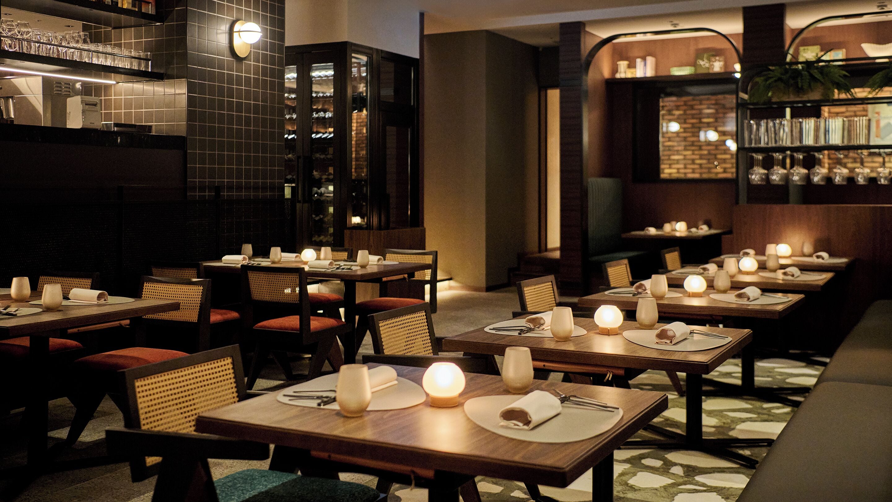 A restaurant with multiple tables set for dining, each with a candle, in a dimly lit interior.