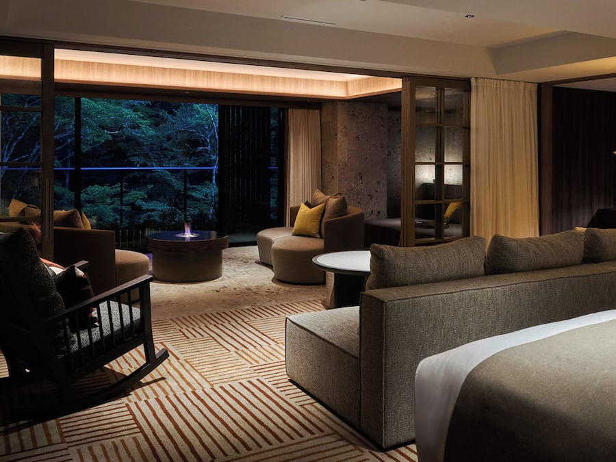 Ein modernes Hotelzimmer mit einem großen Bett, einer Couch, einem Sessel und einem kleinen Tisch. Ein großes Fenster bietet Blick auf Bäume.