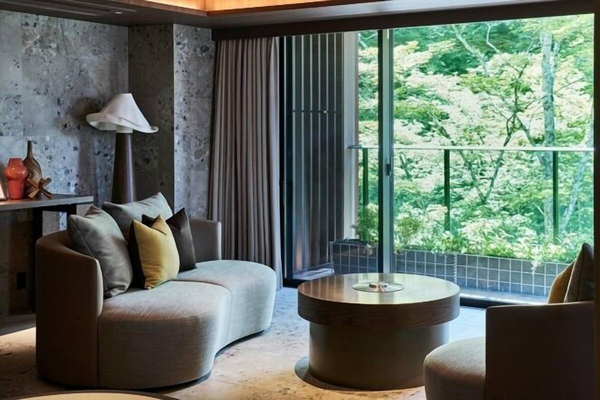 Ein modernes Wohnzimmer mit Sofa, Sesseln, Couchtisch und Lampe. Ein großes Fenster bietet Blick auf Grünflächen.