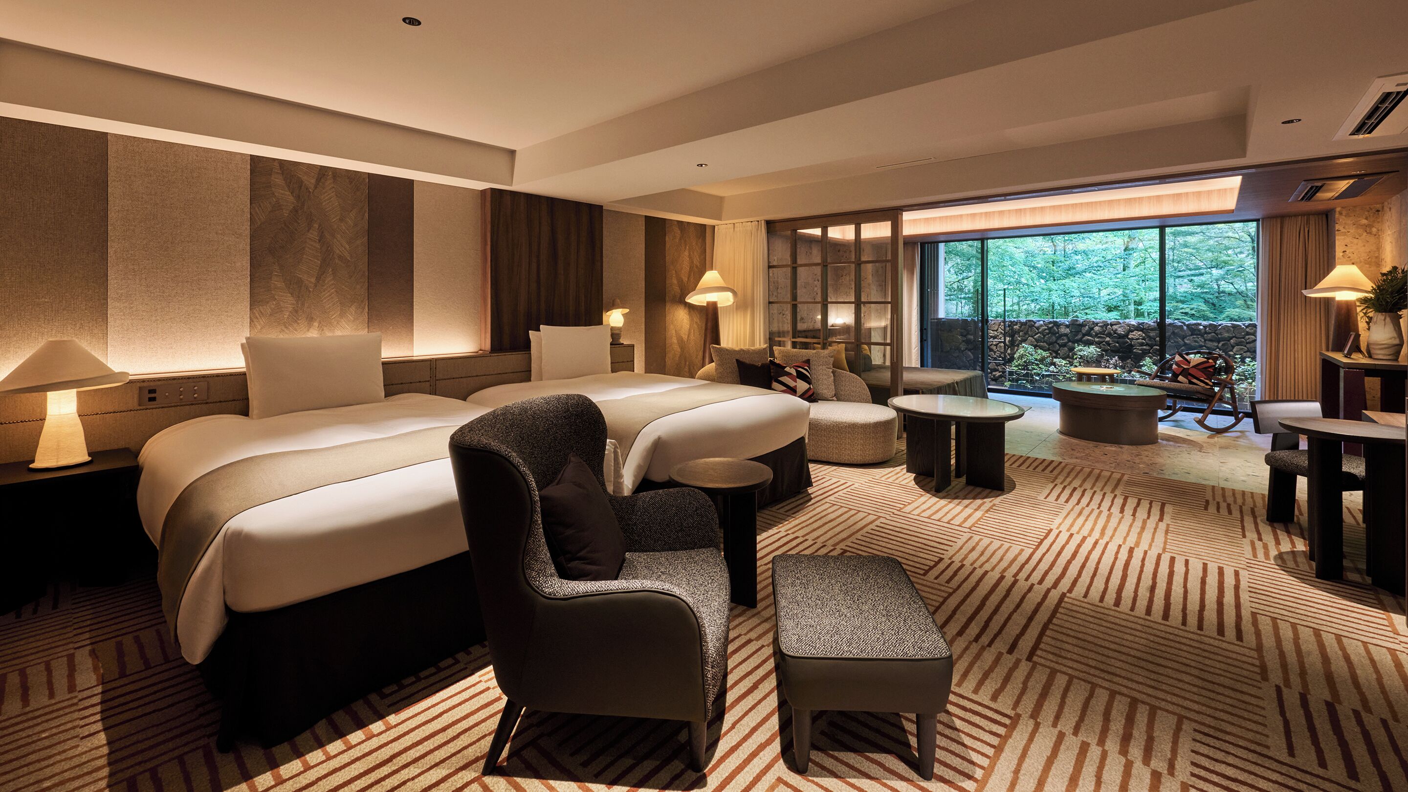 A modern hotel room with a large bed, a sofa, a small table, and a view of the outdoors.