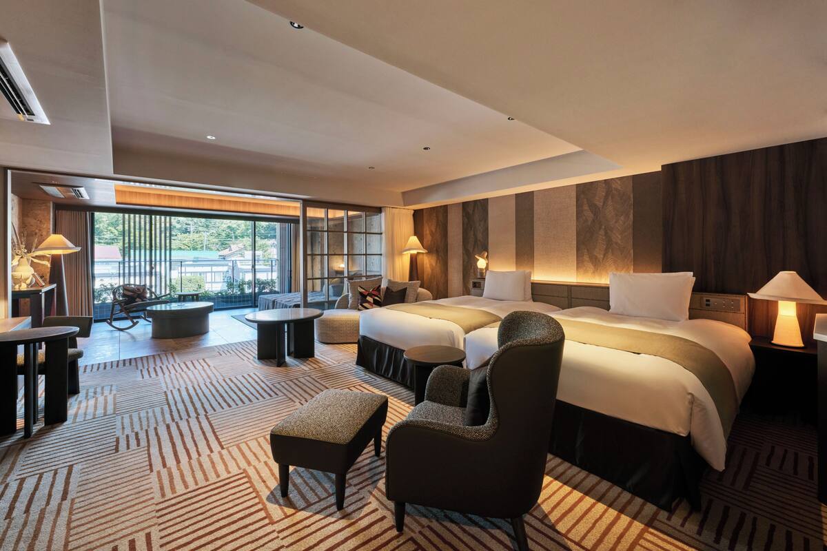 Ein Hotelzimmer mit einem großen Bett, zwei Nachttischen, einem Sofa, einem Sessel, einem kleinen Tisch und einem großen Fenster mit Blick ins Freie.