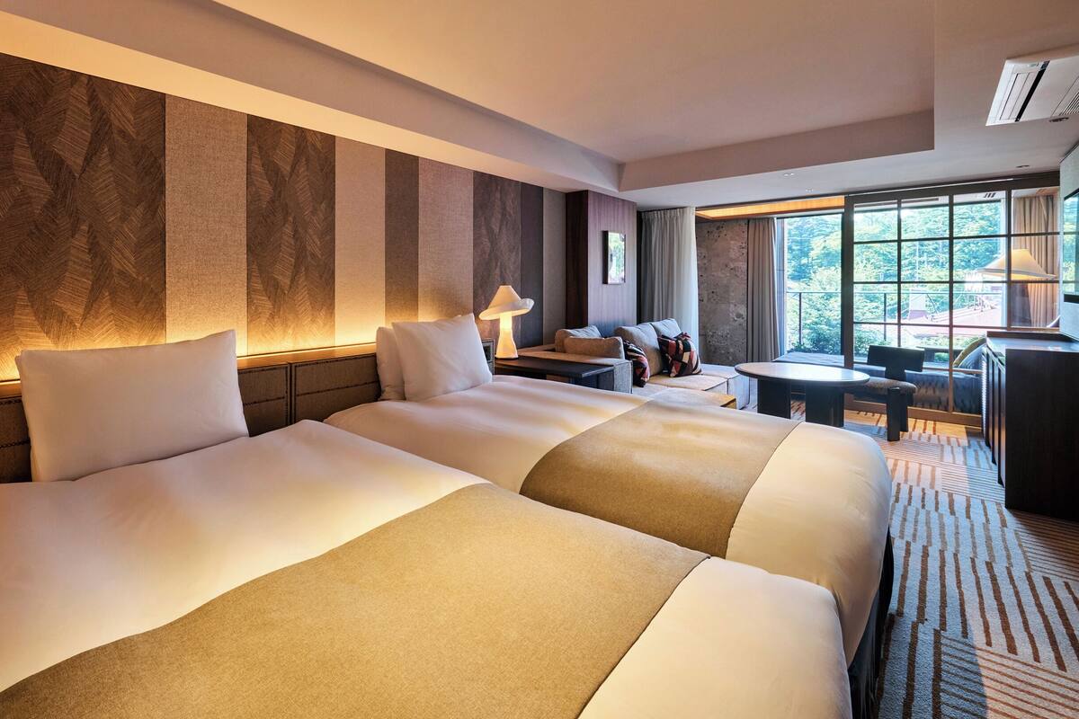 Ein Hotelzimmer mit zwei grossen Betten, einem kleinen Tisch, einer Couch und einem grossen Fenster mit Blick ins Freie.