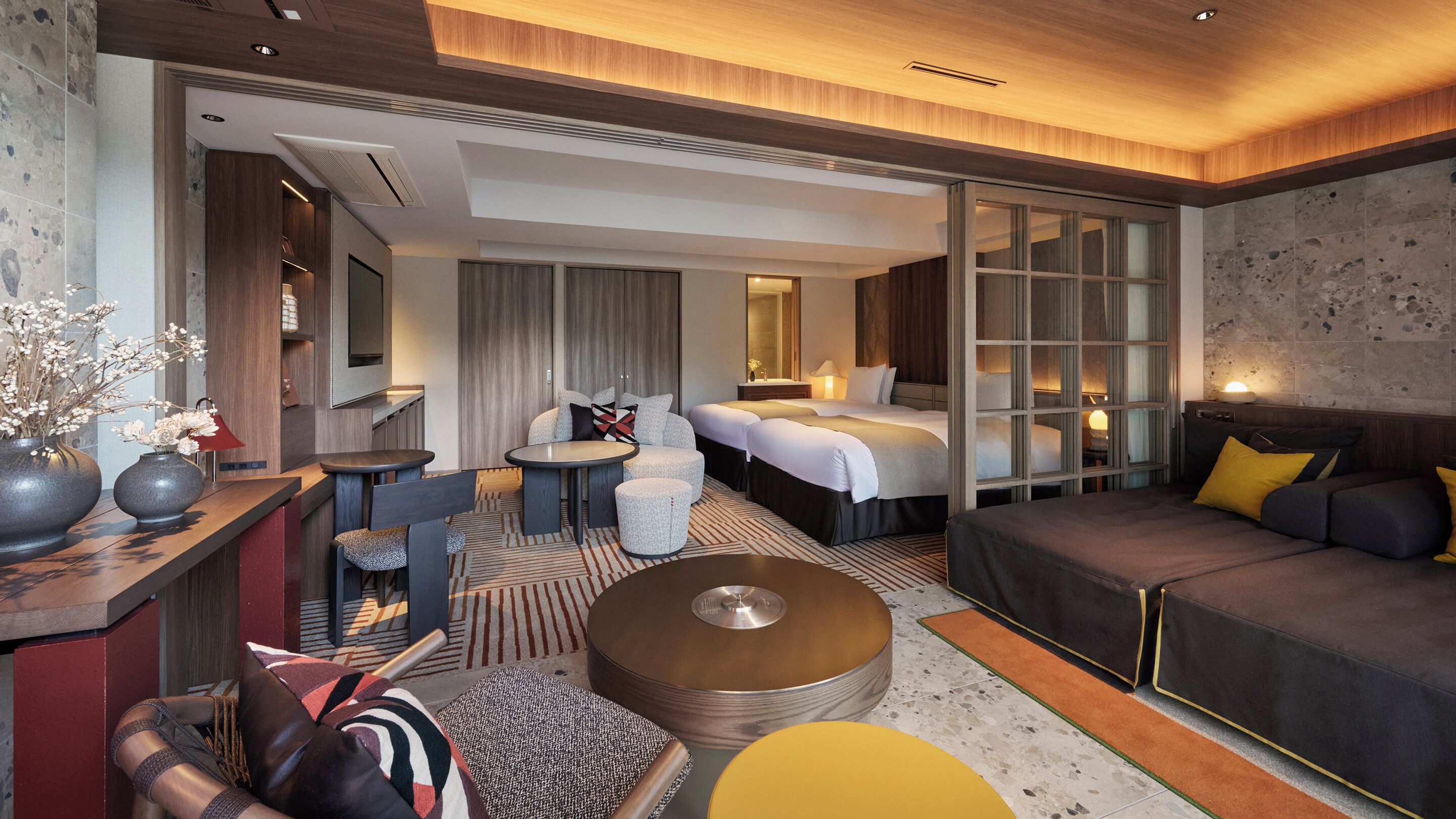 A modern hotel room with a sofa, a round coffee table, a bed with two pillows, a small table with a chair, and a TV on the wall.