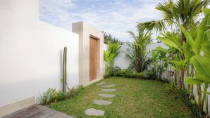 A modern outdoor area with a wooden deck, a stone pathway, and lush greenery including banana plants and a cactus. - Villa Sardegna Ubud (Ubud)