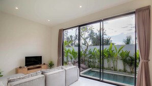 A modern living room with a large TV, grey sofa, and a glass wall overlooking a swimming pool and lush greenery. - Villa Sardegna Ubud (Ubud)
