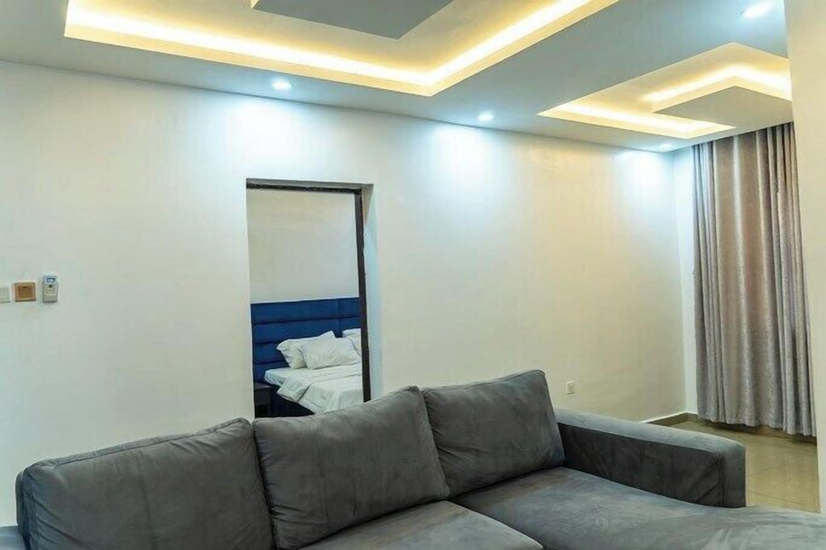 A modern hotel room with a grey sofa, a bed with a blue headboard, and a large mirror.