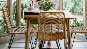 A person seated at a table with a wicker chair, a wicker table, and a window with vertical wooden frames. - Lakubumi Pavilion (Ungasan)