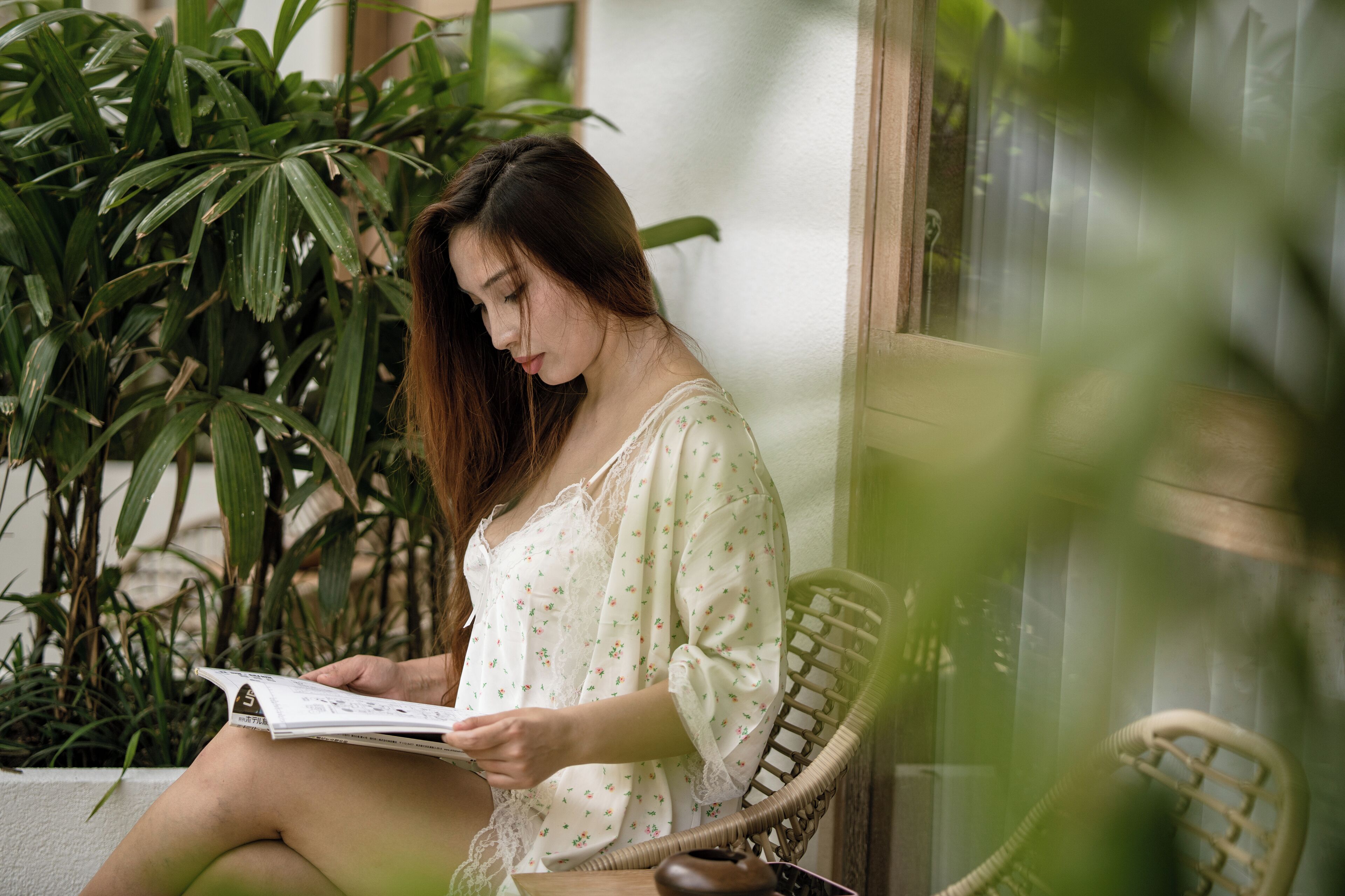 A person sitting on a wicker chair reading a book in a room with large windows and potted plants.