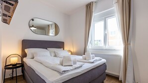 En pænt redt seng med hvide hannelinned og håndklæder, en sengetlampe og et vindue med gardiner.