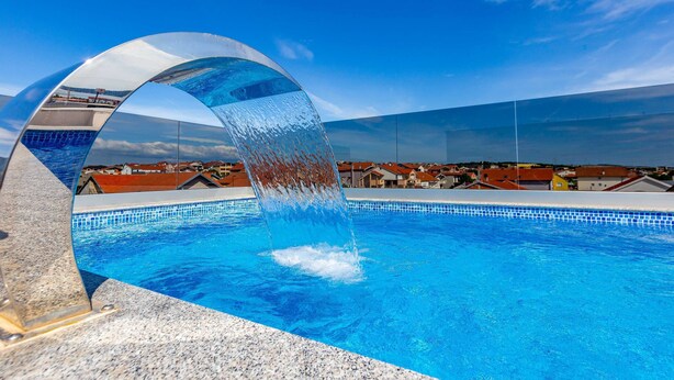 En swimmingpool med klart, blåt vand, en buet glasstruktur og udsigt til huse i baggrunden.
