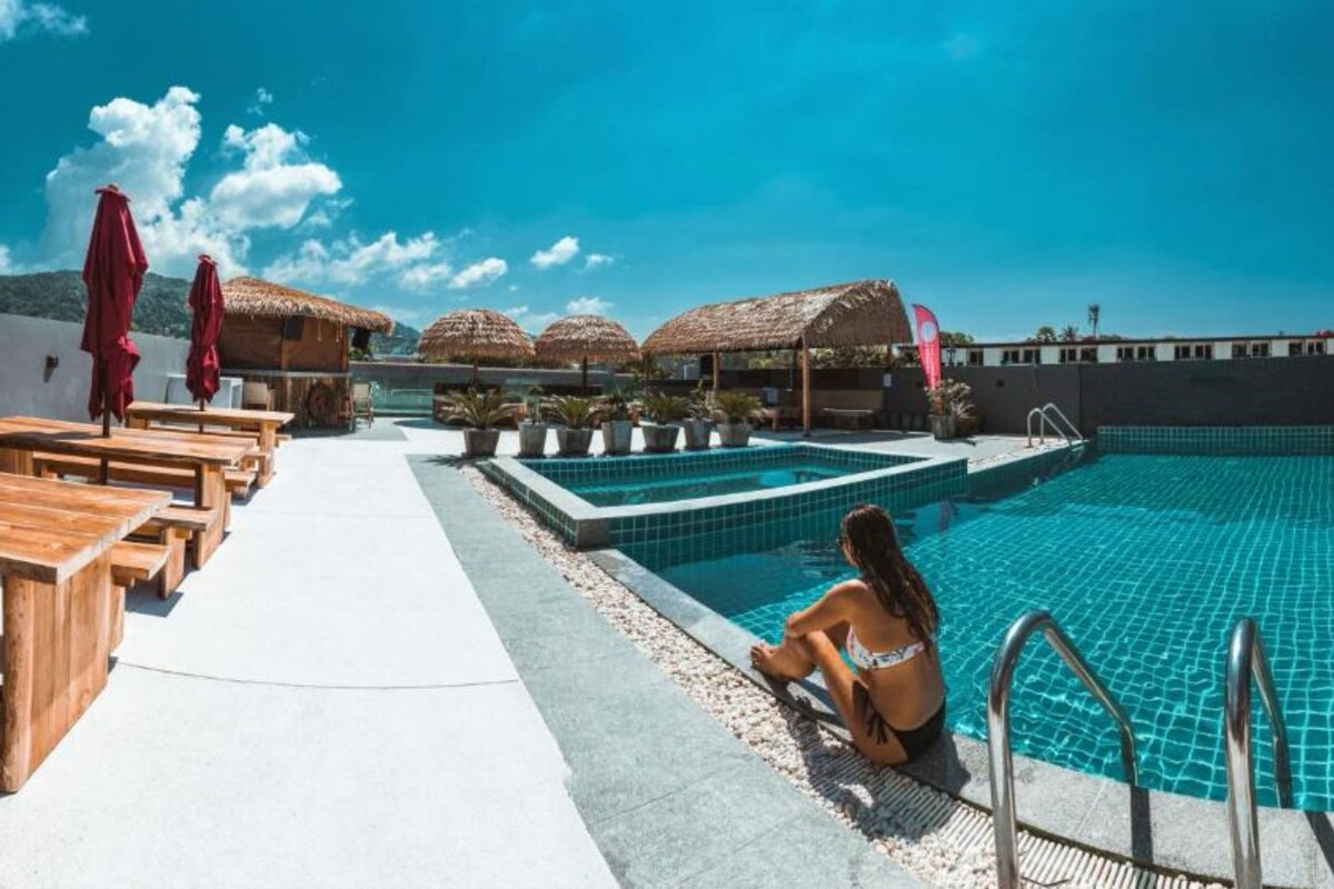 A poolside area with wooden tables and chairs, a swimming pool, and thatched umbrellas.