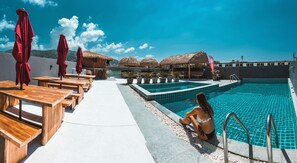 A poolside area with wooden tables and red umbrellas, a clear blue sky, and a woman sitting by the pool. - EBS Hostel Phuket, Kata Beach (Kata)