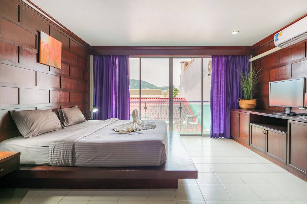A hotel room with a bed, a TV, a balcony with purple curtains, and a view of buildings and mountains.