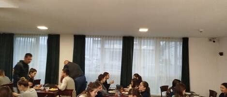 Un comedor con mesas y sillas, gente comiendo y una ventana con cortinas.