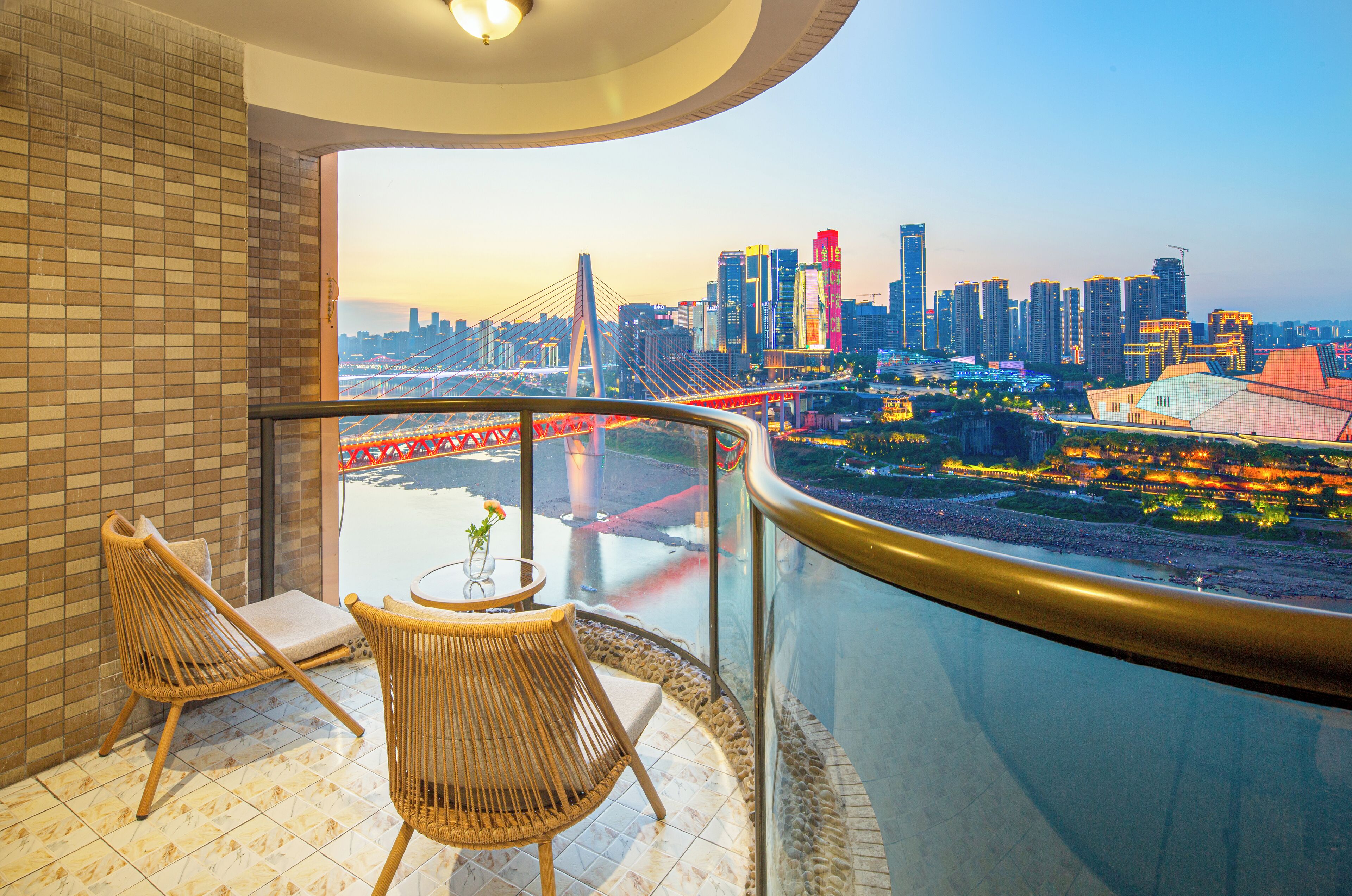 A balcony with wicker chairs and a table, overlooking a cityscape with a bridge and high-rise buildings.