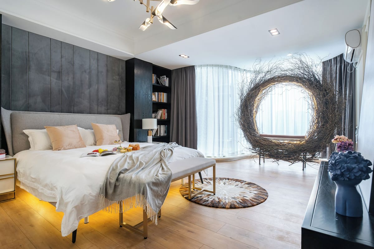 A modern bedroom with a large bed, a bedside table, a bookshelf, a round mirror, and a window with curtains.