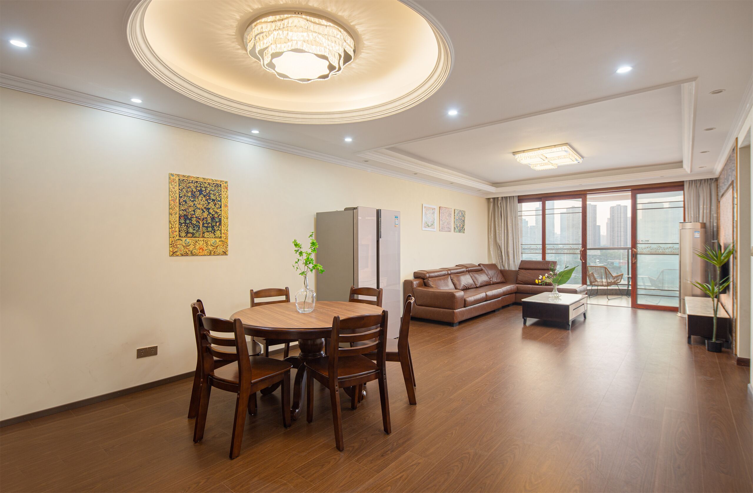 A spacious living room with a dining area, a brown leather sofa, a chandelier, and a large window with a city view.