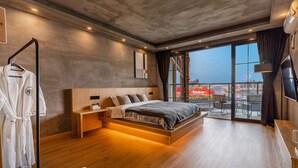 A modern hotel room with a large bed, a wooden bedside table, a robe hanging on a rack, and a city view through large windows. -  Nuanyu Apartment with River View & Drone Show View (Chongqing)