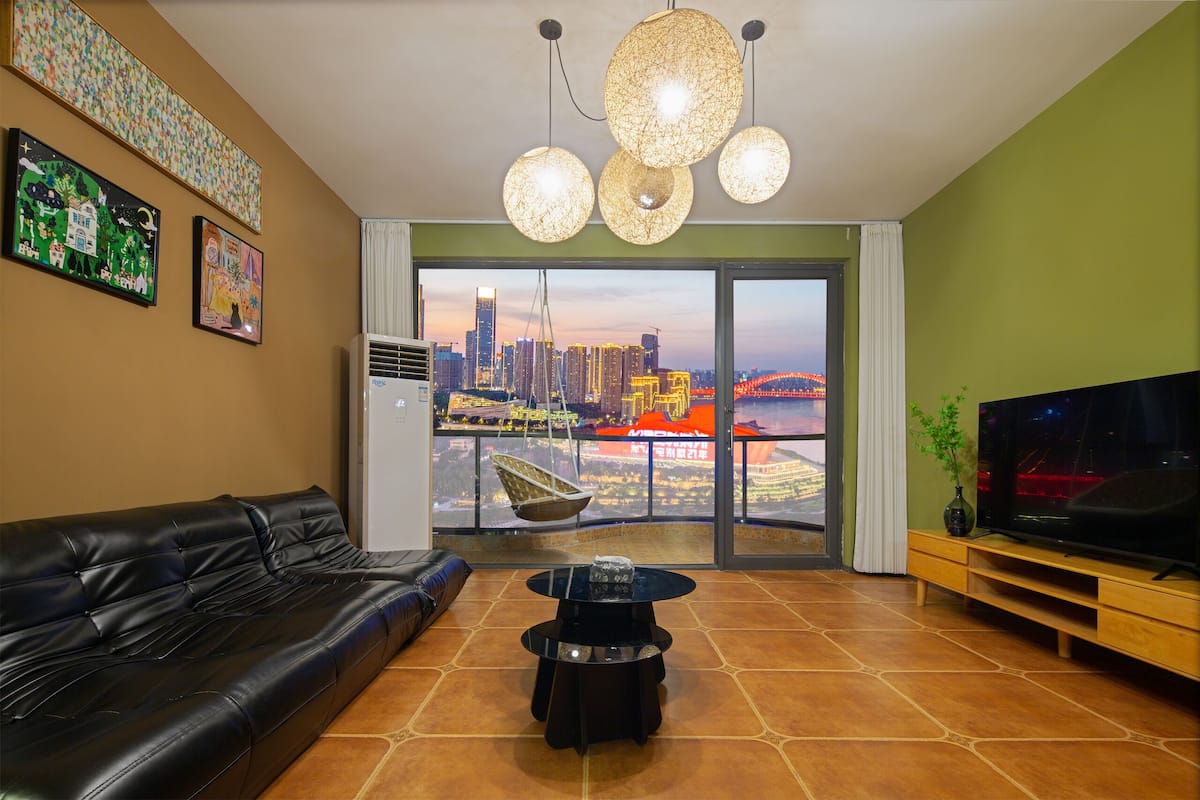 A living room with a black leather sofa, a round coffee table, and a television on a wooden stand. There are two hanging spherical light fixtures and a large window with a cityscape view.