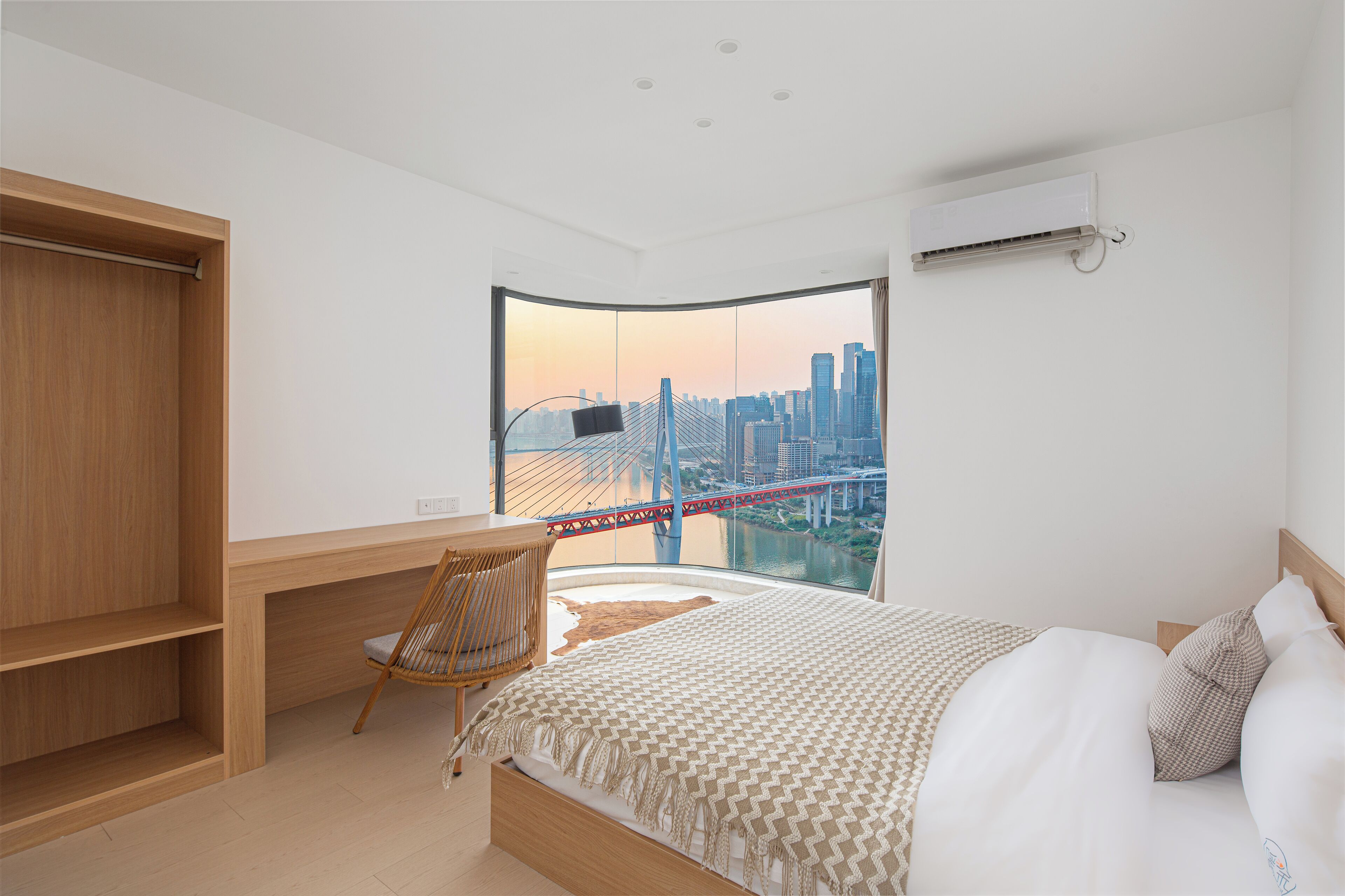 A modern bedroom with a large bed, a desk, and a chair, featuring a cityscape view through a large window.