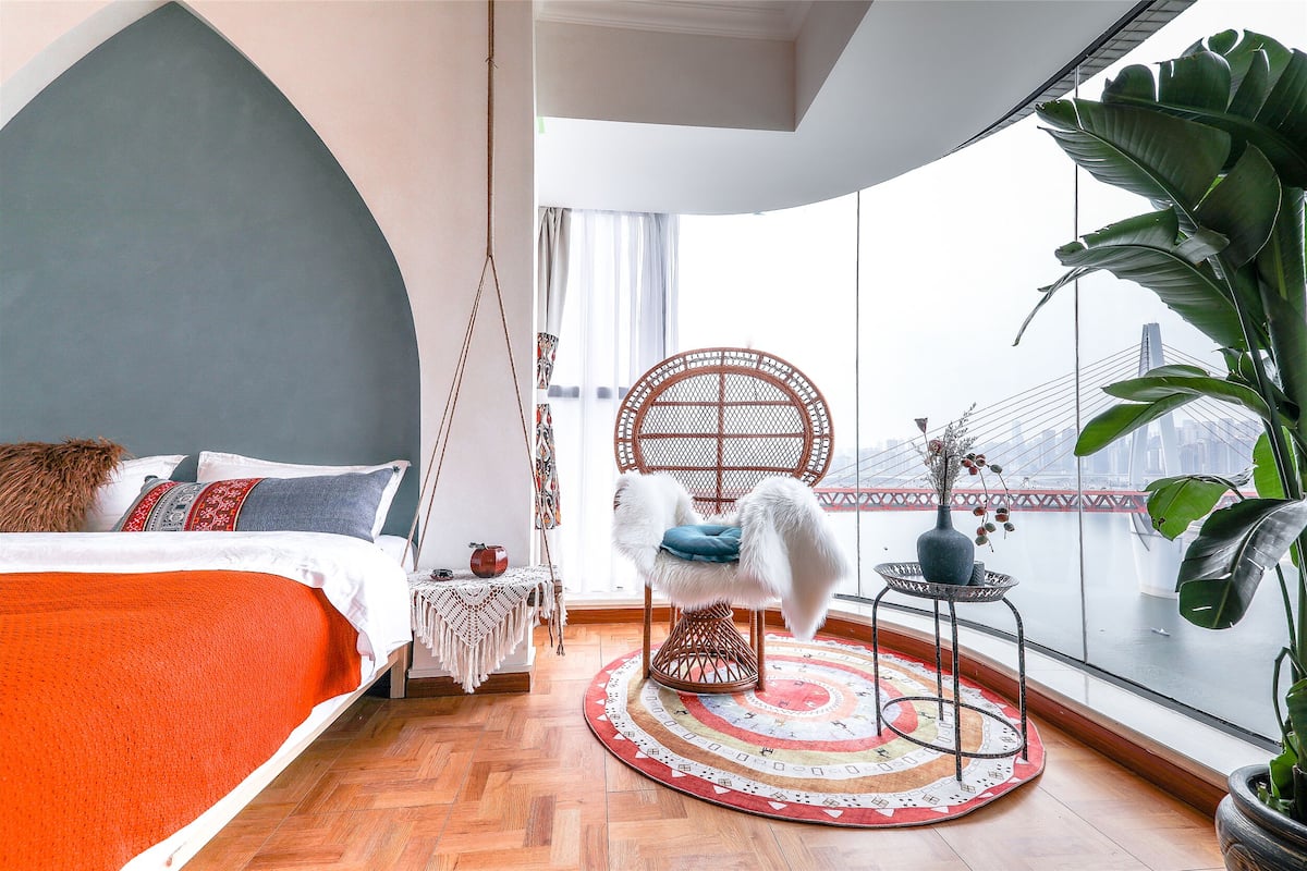 A modern bedroom with a large bed, a round wicker chair, a small table, and a view of a cityscape through large windows.