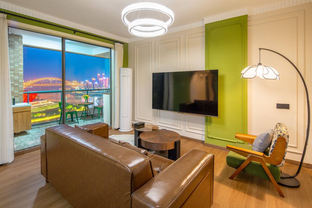A modern living room with a large flat-screen TV, leather sofas, a round coffee table, and a view of a cityscape through floor-to-ceiling windows.