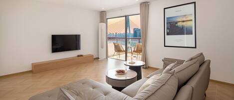 A modern living room with a large sofa, a flat-screen TV, a dining table with chairs, and a large window with a city view.