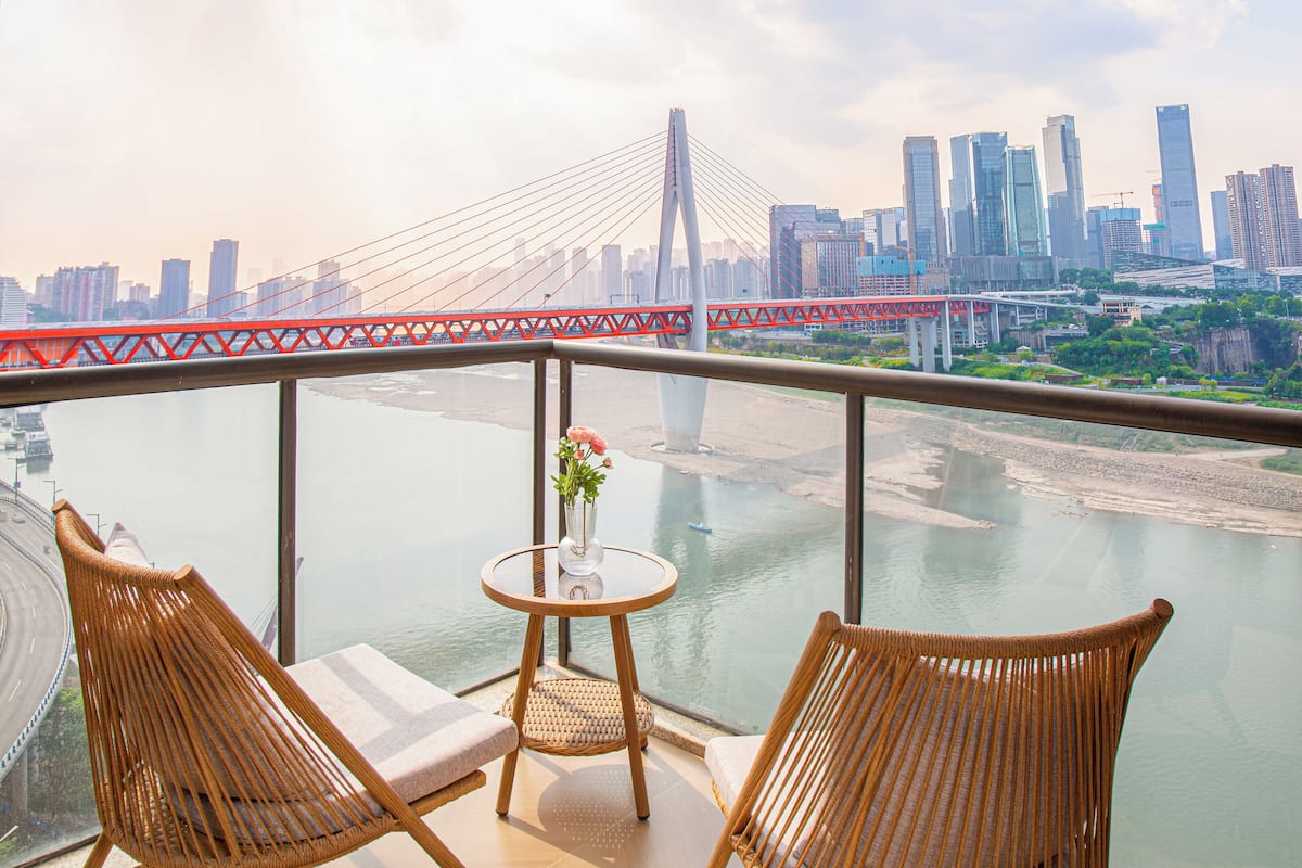A balcony with wicker chairs and a small table, overlooking a river and a cityscape with a prominent bridge.