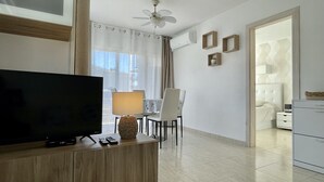 A modern hotel room with a TV on a wooden desk, a dining area with chairs and a table, and a bedroom with a bed and dresser.