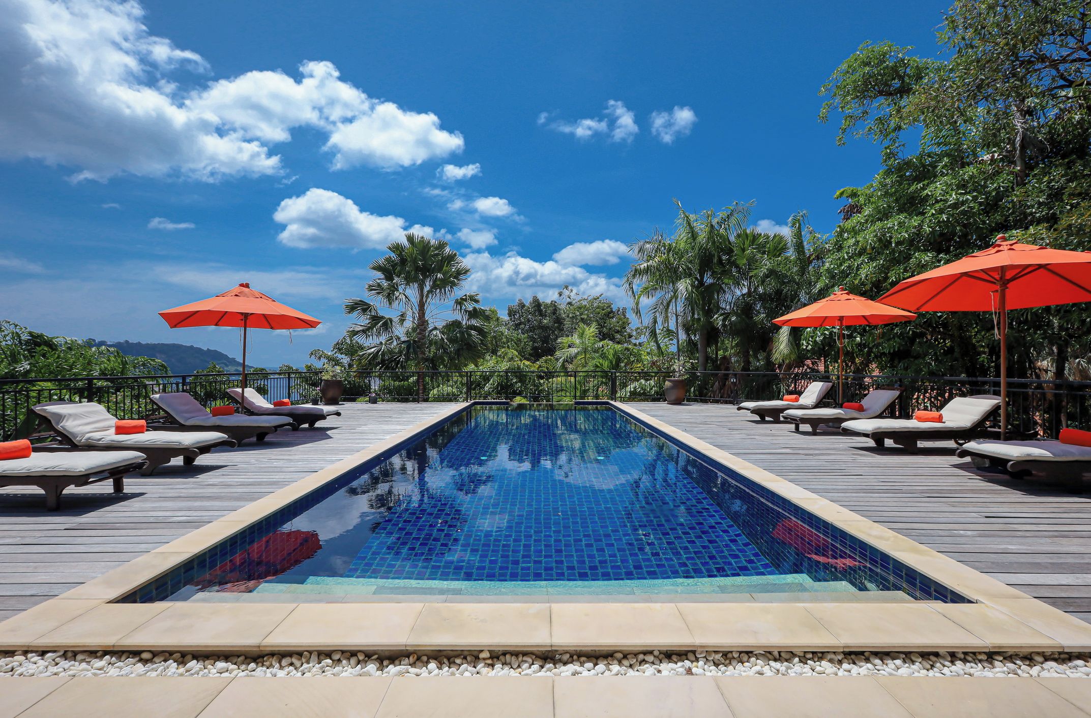 A rectangular pool with tiled blue surface, surrounded by lounge chairs with orange cushions, and red umbrellas.