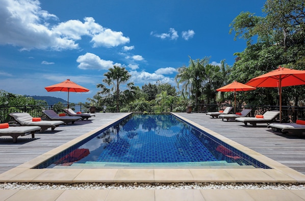 A rectangular pool with tiled blue surface, surrounded by lounge chairs with orange cushions, and red umbrellas.
