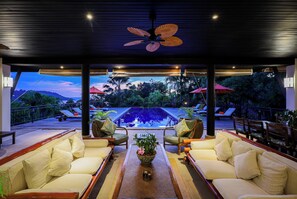 Un salon au bord de la piscine, doté d’un ventilateur de plafond, de sièges confortables et offrant une vue sur la piscine et un paysage tropical.