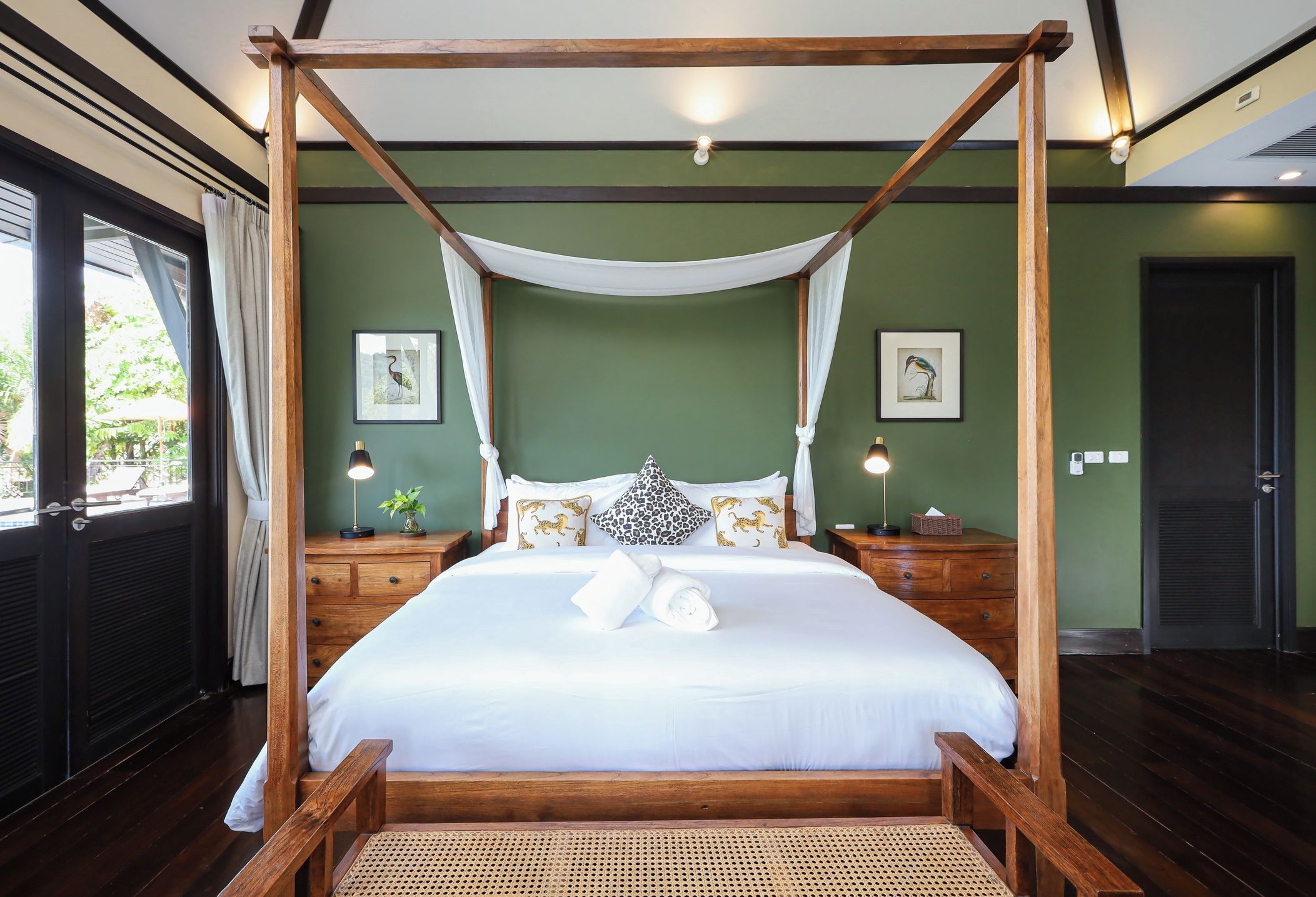 A four-poster bed with white linens, wooden headboard, and a canopy. There are two bedside tables with lamps, a nightstand, and a wall-mounted shelf with a plant.