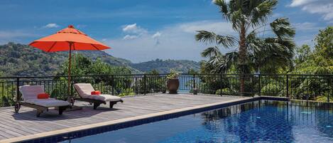 A pool area with two lounge chairs, an orange umbrella, and a view of hills.
