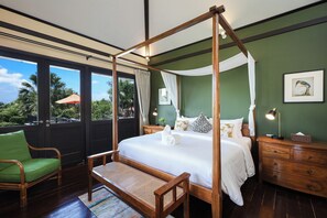 A four-poster bed with white linens, a wooden nightstand with a lamp, a green chair, and a view of palm trees through large windows.