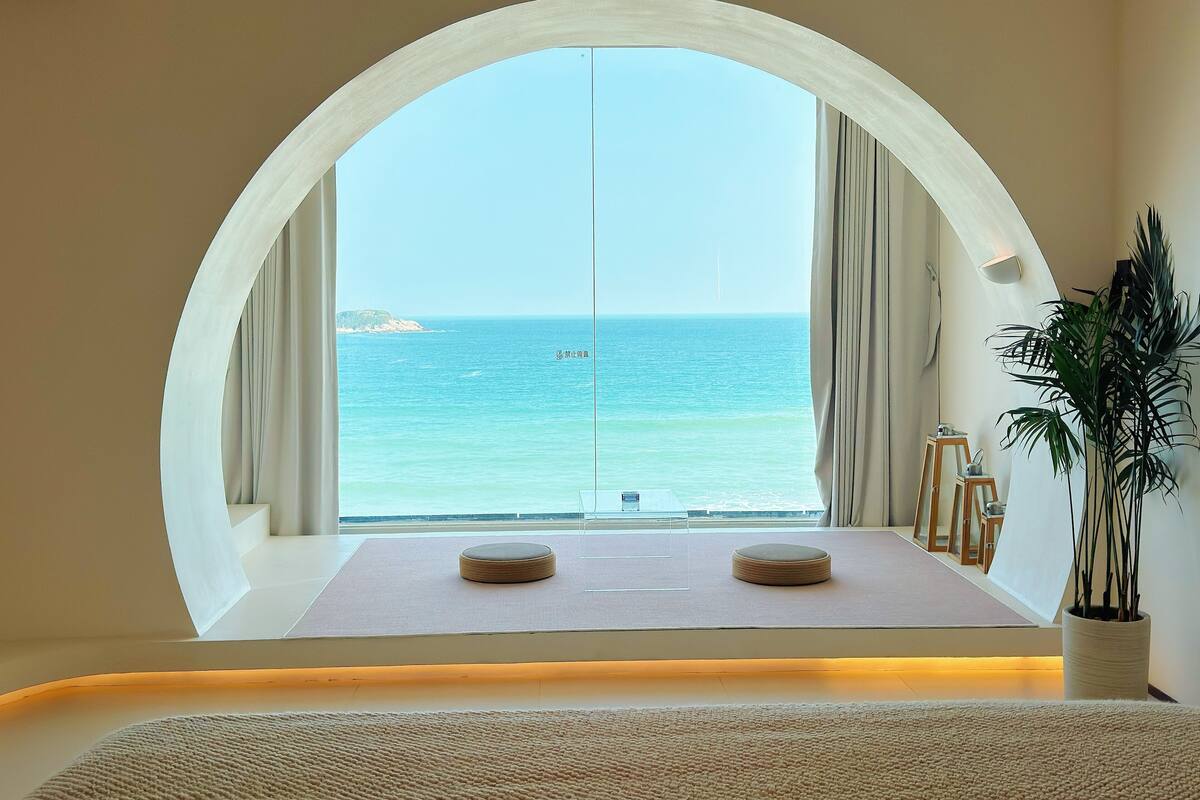 A room with a large arched window overlooking the sea, a small table with a cushion, and a potted plant.