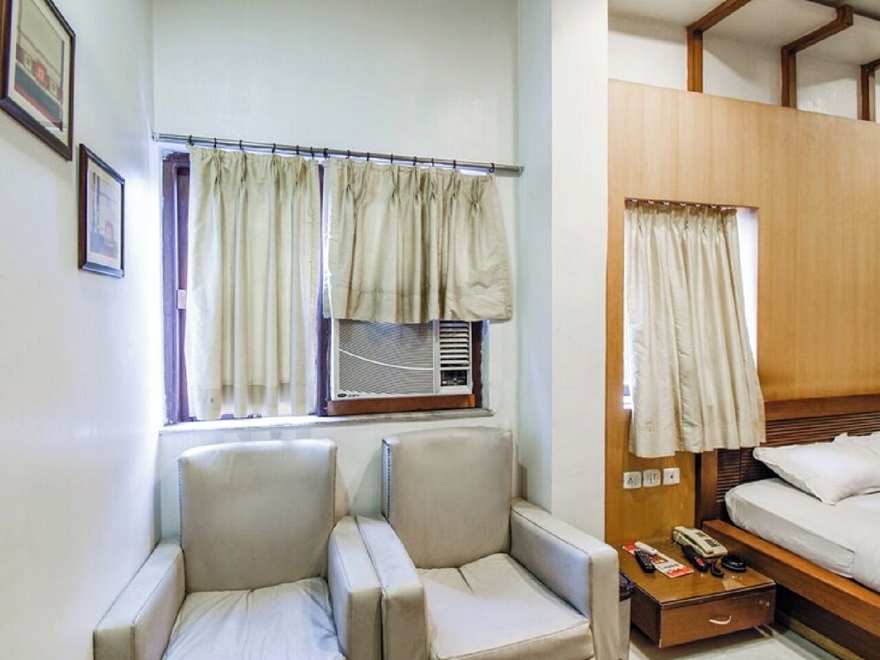 Quarto compacto com duas poltronas acolchoadas, uma mesa pequena com controle remoto e uma cama com cabeceira de madeira.