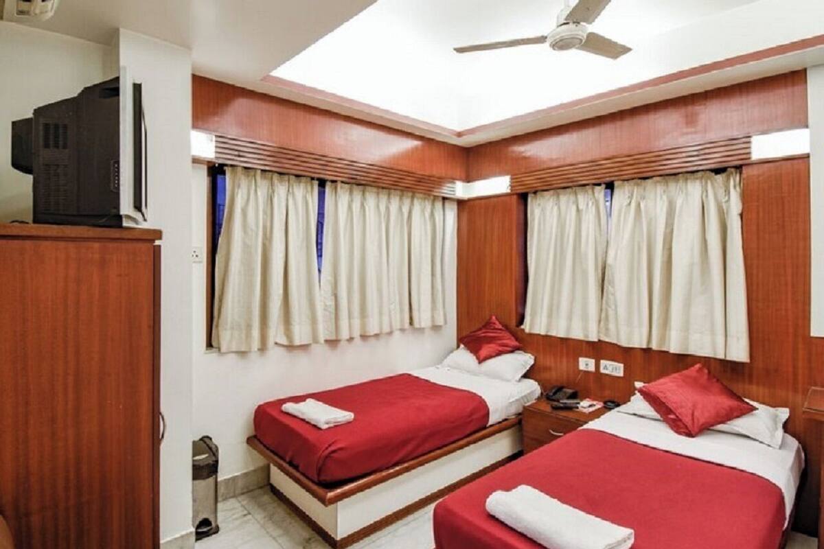 A hotel room with two single beds, wooden headboards, a ceiling fan, and a television on a stand.